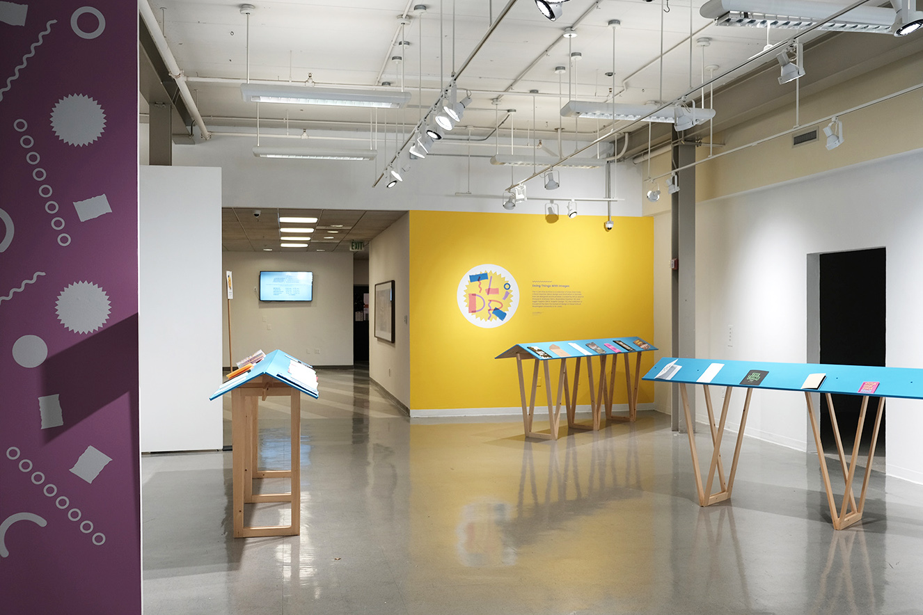 an image of an exhbiition space with a yellow wall, blue readers with zines on them, and little purple wall