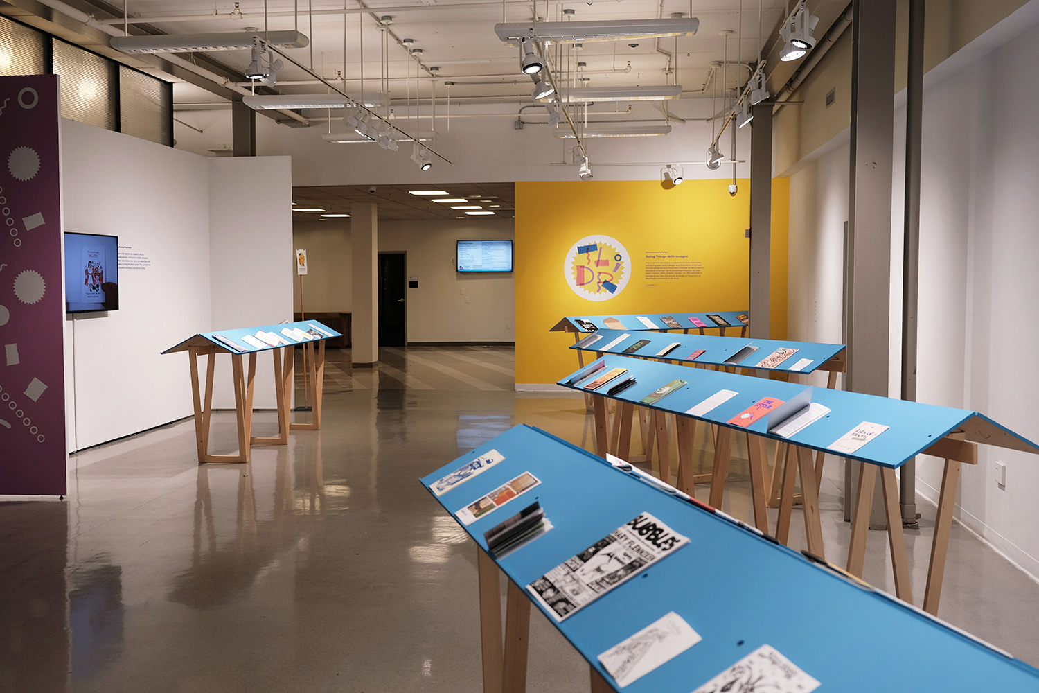 an image of an exhibition with a yellow wall, a purple wall, and blue readers that have zines on them