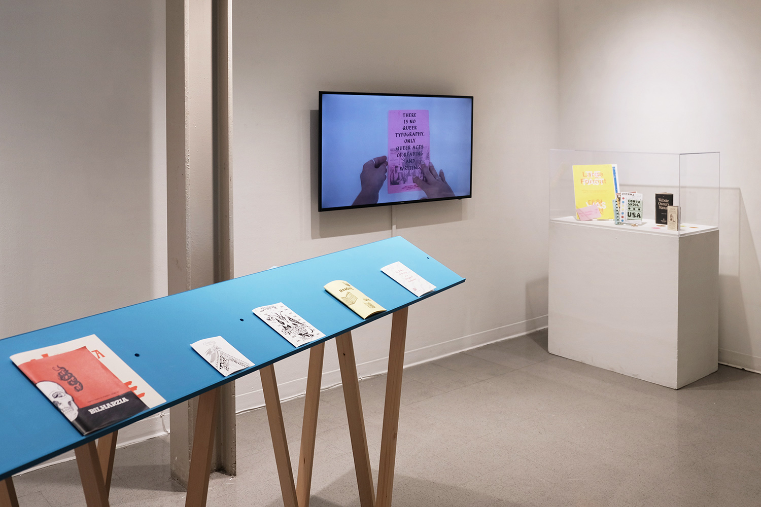 an image of an exhibition space with blue readers that have zines on them, a monitor displaying a zine, and a vitrine holding zines