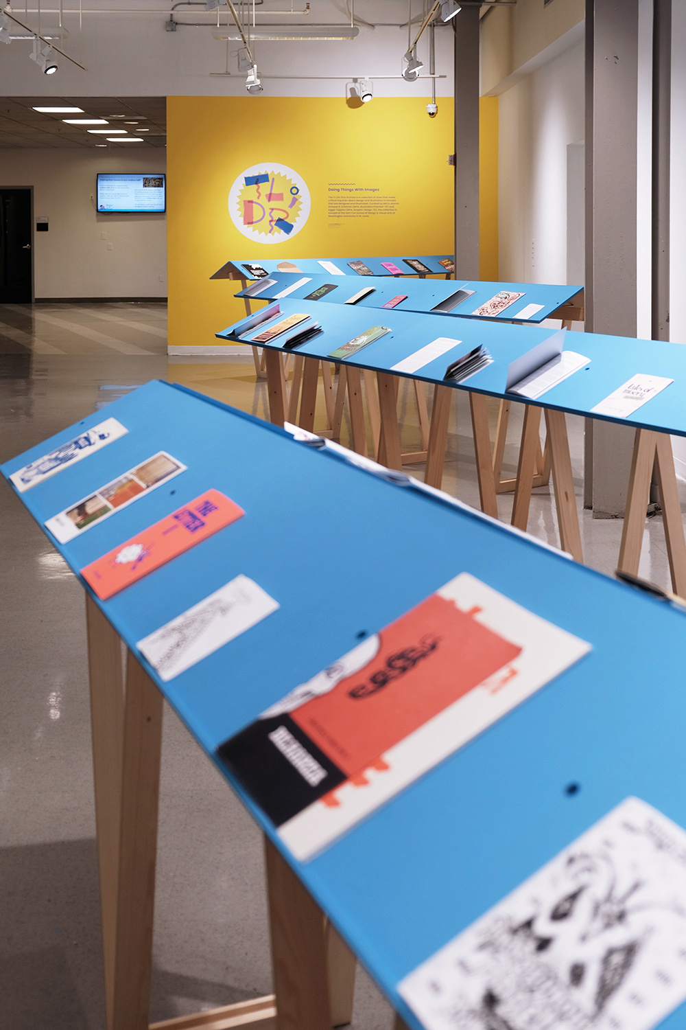 an image of an exhbition space with a yellow wall, blue readers with zines on them, and little purple wall